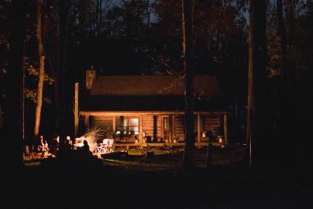 Symbolbild: Alte Berghütte im Wald bei Dunkelheit. Licht kommt nur von einem Lagerfeuer vor der Tür.